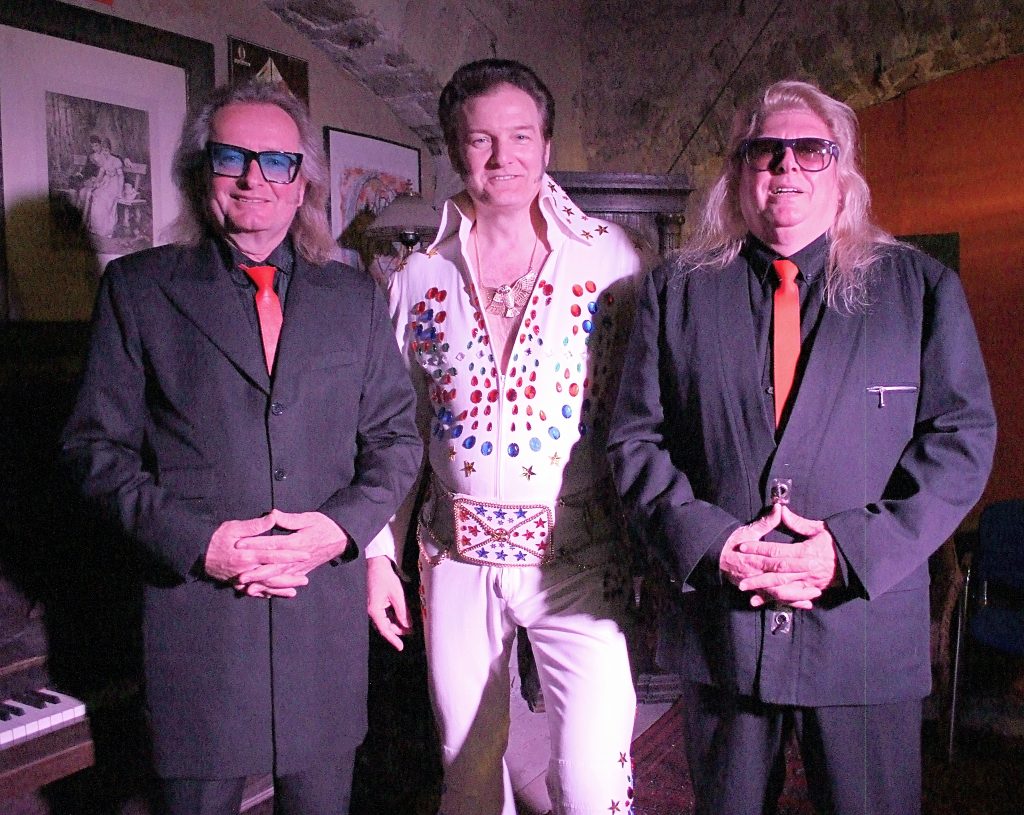 Links und rechts stehen 2 Männer der Boogie Band in schwarzen Anzügen mit roten Krawatten. Dazwischen steht der Elvis Imitator.
