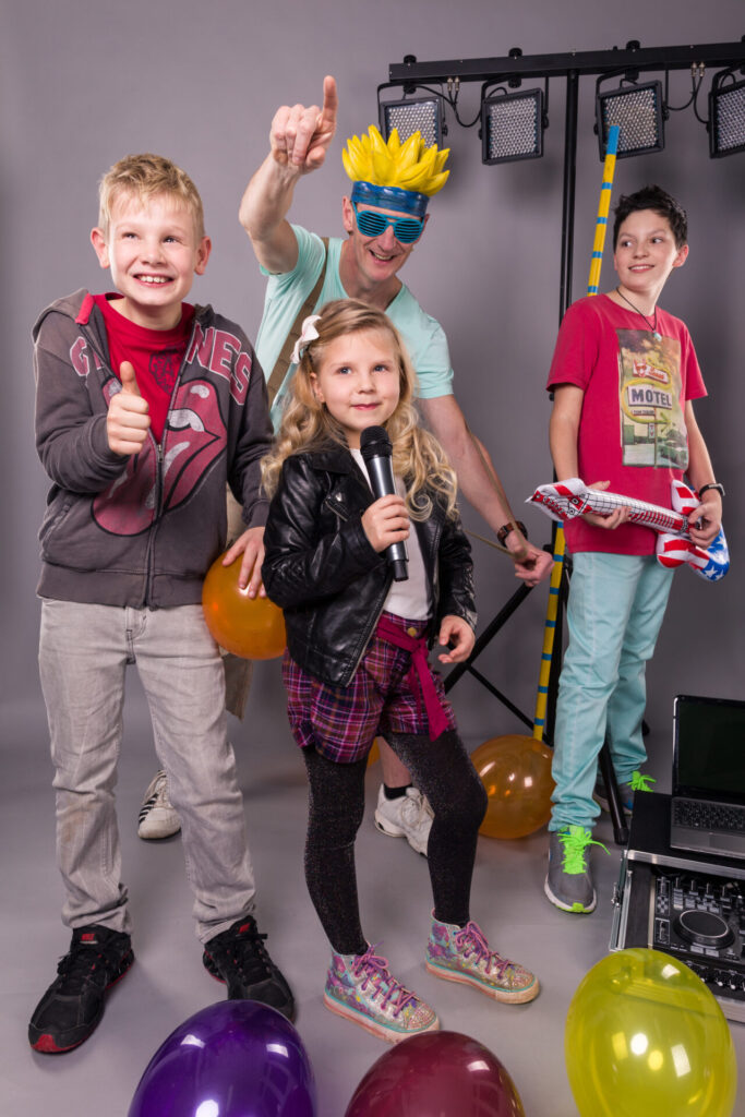 Der DJ und drei Kinder, die ein Mikrofon und eine Luftgitarre halten. Im Hintergrund sind Luftballons.
